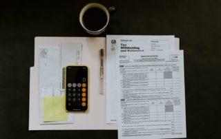 foto de uma pasta aberta com papéis em cima, um celular com a calculadora aberta e um café acima da imagem para simular a realização de um balanço patrimonial